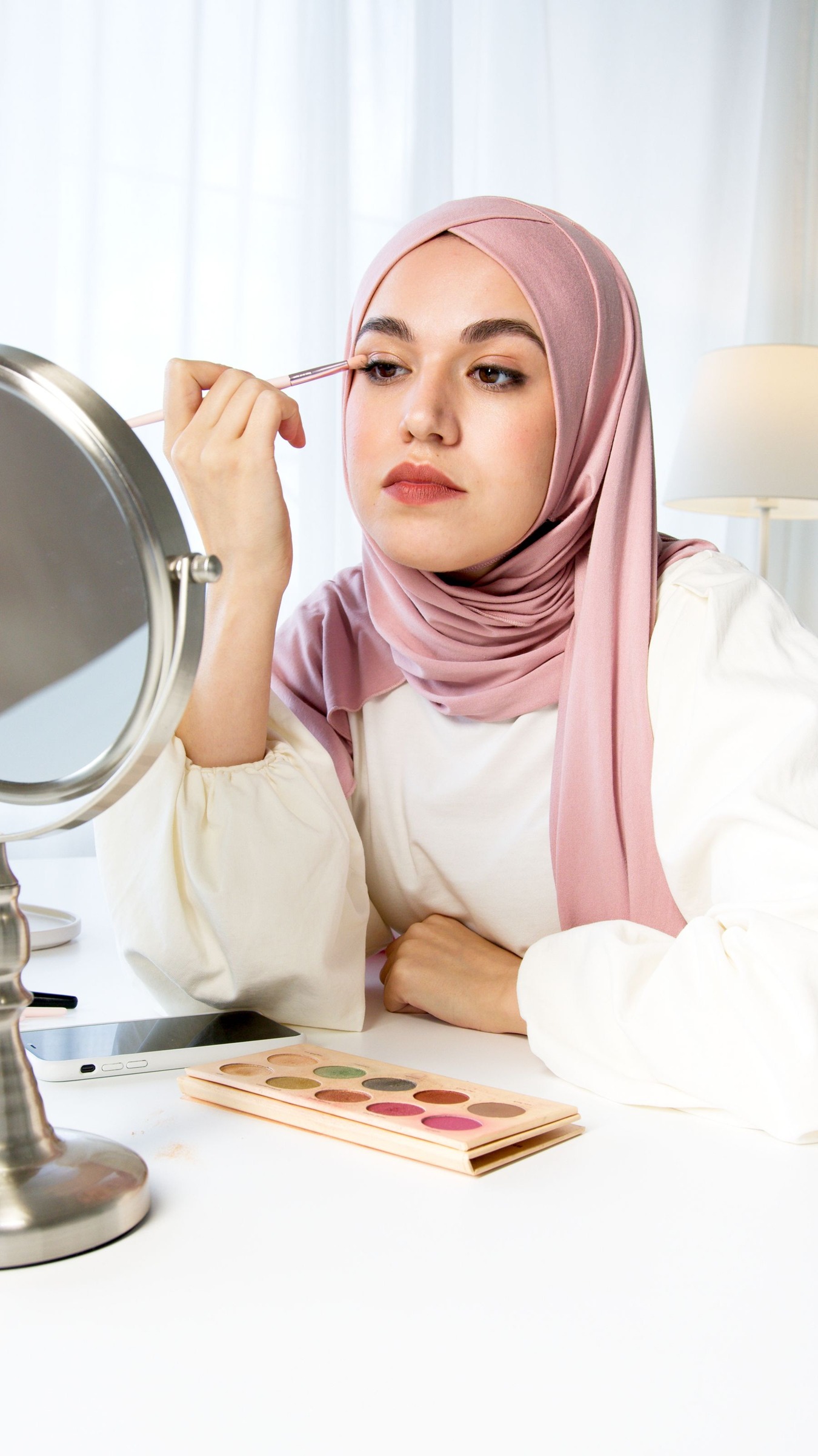 Woman in a pink hijab applying eyeshadow while looking in a mirror