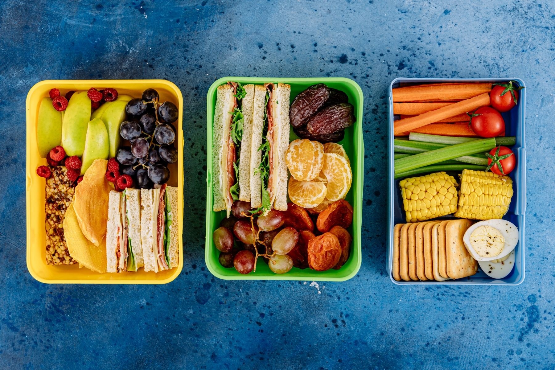 Three lunch boxes with sandwiches, fruit and vegetables on a blue background