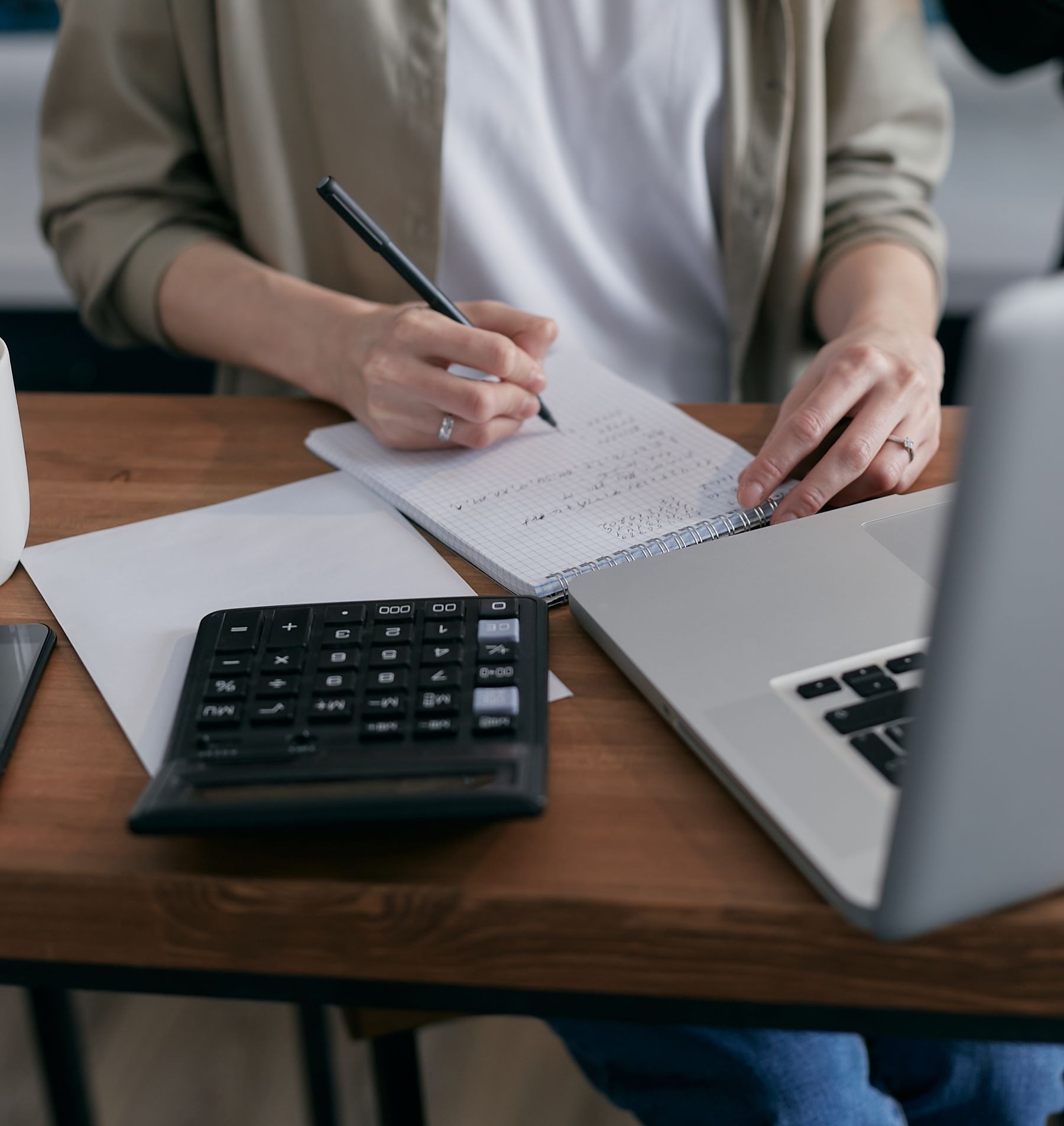 Woman writing budget calculations in a notebook beside a calculator and laptop