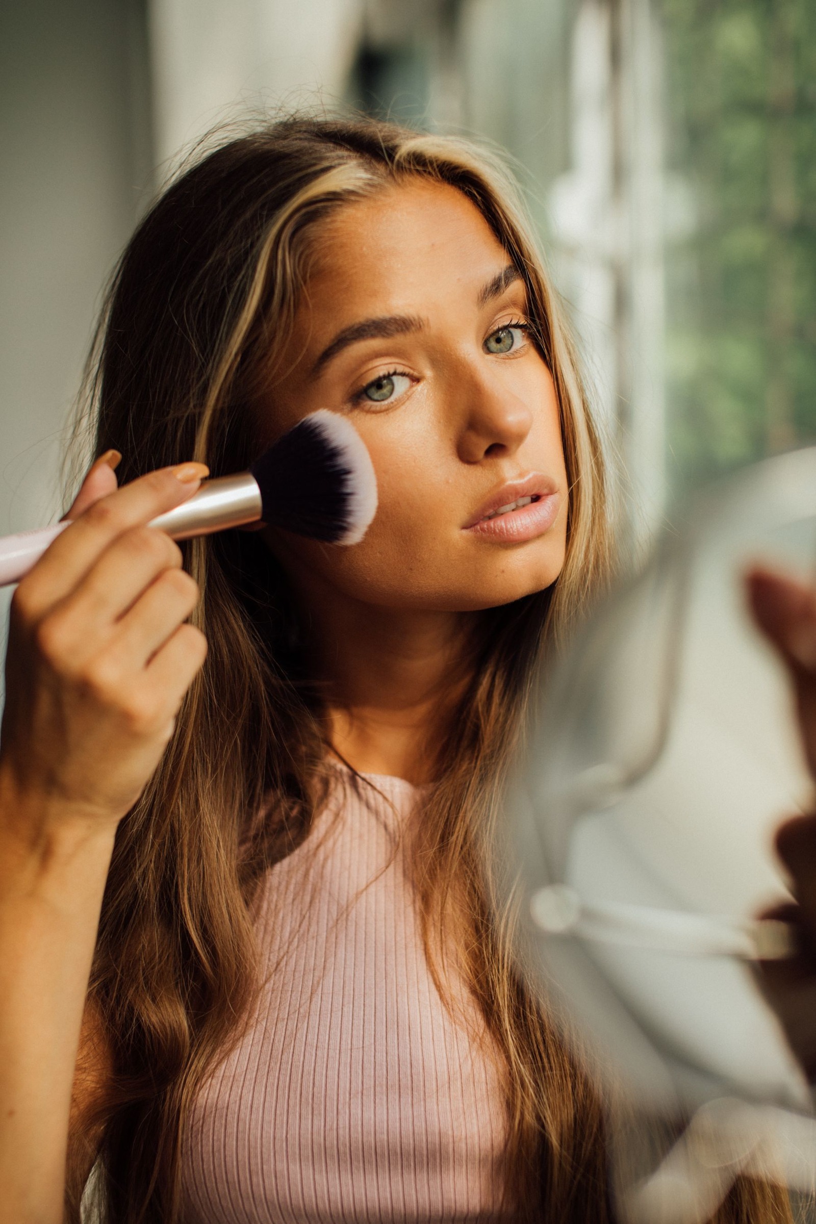 Woman using a cheek brush to apply blush beside a window